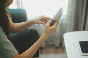 Happy Asian teen girl holding pad computer gadget using digital tablet technology sitting on the couch at home. Smiling young woman using apps, shopping online, reading news, browsing internet on sofa photo