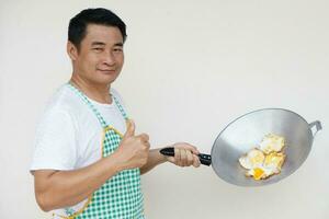 Handsome Asian man is cooking fried eggs, wears white shirt and apron, holds frying pan and ladle spatula , thumbs up. Concept, love cooking. Kitchen lifestyle. Look at camera. Copy space for text photo