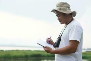 asiático hombre ecologista es topografia naturaleza a el lago, escribir en papel cuaderno. concepto, naturaleza exploración. ecología estudiar. pasatiempo actividad, estilo de vida. hombre explorar ambiente foto