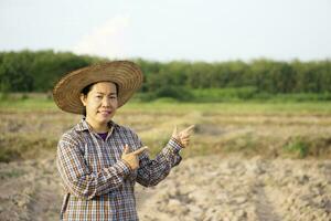 Asian woman farmer wears hat, plaid shirt, point fingers and smile, stands at agricultuture land, feels confident. Concept , happy farmer. Agriculture occupation. Copy space for adding advertisement. photo