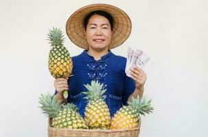 Asian woman gardener wear hat, blue shirt, holds pineapple fruit and Thai banknotes money. Concept , Agriculture crop in Thailand. Farmer satisfied, get income. Organic crops. Seasonal fruits. photo