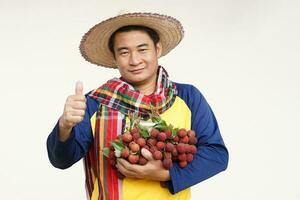 Handsome Asian man farmer wears hat, yellow shirt, holds litchi fruits. Thumbs up. Concept, agriculture occupation. Thai farmers grow organic litchis as an export product of Thailand. photo