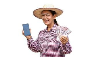 Happy senior woman farmer wears hat, plaid shirt, holds Thai banknotes money and mobile phone, isolated on white background. Concept, Get profit, income from doing agriculture. Use online apps. photo