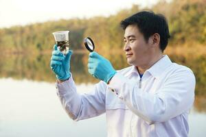 Asian man ecologist holds magnifying glass to inspect water in transparent glass from the lake. Concept, explore, analysis water quality and creature from natural source. Ecology field research. photo