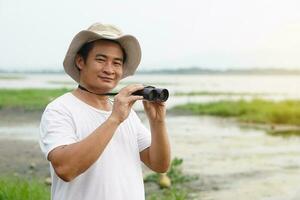 Handsome Asian man ecologist is surveying nature at the lake, hold binoculars.Concept, nature exploration. Concept, nature exploration. Ecology study.  Pastime activity, lifestyle. Explore environment photo