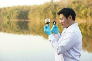 Asian man ecologist holds magnifying glass to inspect water in transparent glass from the lake. Concept, explore, analysis water quality and creature from natural source. Ecology field research. photo
