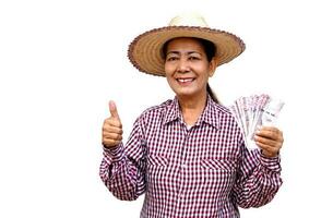 Happy senior woman farmer wears hat, plaid shirt and holds Thai banknotes money. Thumbs up, isolated on white background. Concept, Farmer happy to get profit, income from doing agriculture. photo