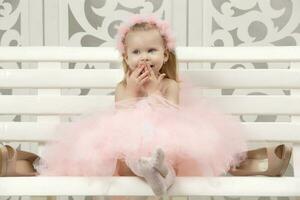 Two-year-old child. A beautiful little girl in a smart pink dress sits on a wooden bench and laughs. A cute baby. photo