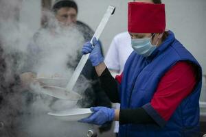 noviembre 13, 2020. bielorrusia, gomil región. Pensilvania Cocinando calle comida durante el coronavirus panademia un cocinero en un médico máscara con un grande cucharón vierte sopa. foto