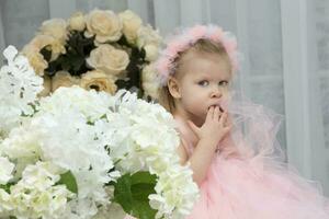 un pequeño niña en un elegante vestir con un triste expresión soportes entre el flores gracioso niño. foto