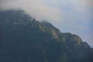 Dark atmospheric surreal landscape with a dark rocky mountain peak in low clouds in a gray cloudy sky. A gray low cloud on a high peak. High black rock in low clouds. Surreal gloomy mountains. photo