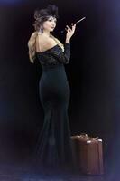 A beautiful girl dressed in the style of the 20-30s of the last century with a cigar and a suitcase on a dark background. Vintage retro girl. photo