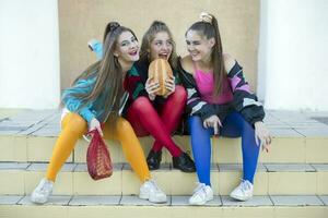 Three pretty girls dressed in the style of the nineties are sitting on the steps and sharing a roll. photo