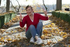 A happy middle-aged woman sits on a footpath in an autumn park and shows a class sign with her fingers. photo