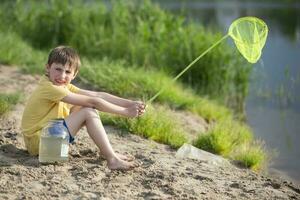 A little boy with a fishing net and a jar sits on the banks of a river or lake. photo
