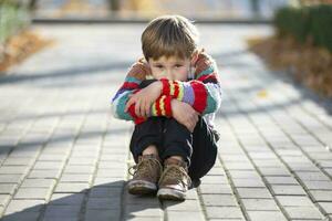 triste pequeño chico se sienta en el callejón en el parque. el niño poner su cabeza en su rodilla. foto