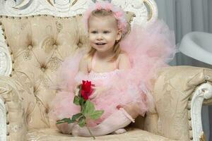 A little girl in an elegant pink dress with a red rose sits in a chair. photo