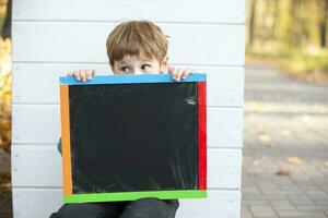 A little boy sits in the park near the wall and holds an empty black board for the inscription. photo