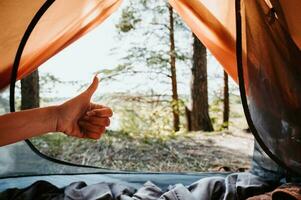 Camping. View from the tent. A woman's hand shows the Cool sign against the background of trees and water. Outdoor recreation. photo