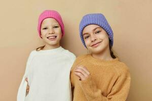 two little girls in sweaters and hats hug friendship beige background photo