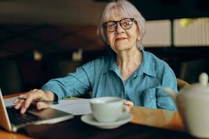 Beautiful mature senior woman with glasses sits at a table in front of a laptop unaltered photo