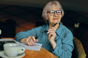 elderly woman with glasses sits at a table in front of a laptop unaltered photo
