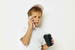 pequeño chico en un blanco camiseta gorra con un teléfono en un vaso con un bebida ligero antecedentes inalterado foto