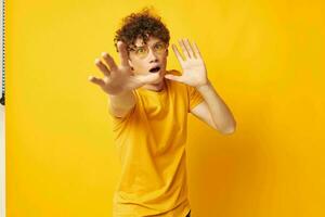 Young curly-haired man yellow t-shirt glasses fashion hand gestures yellow background unaltered photo