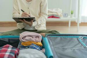 viaje bolsa. hembra son comprobación el detalle y cantidad de artículos usado durante el viaje por Lista de Verificación. joven mujer son preparar ropa y personal pertenencias. paquete tu equipaje antes de viajar. foto