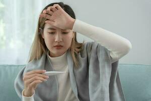 Young Asian woman having high fever while checking body temperature, female sneezing and runny nose with seasonal influenza, allergic, digital thermometer, virus, coronavirus, illness, respiratory photo