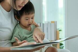 la feliz madre asiática se relaja y lee un libro con el tiempo del bebé juntos en casa. los padres se sientan en el sofá con su hija y leen una historia. aprender el desarrollo, el cuidado de los niños, la risa, la educación, la narración de cuentos, la práctica. foto