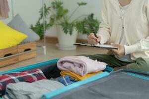 viaje bolsa. hembra son comprobación el detalle y cantidad de artículos usado durante el viaje por Lista de Verificación. joven mujer son preparar ropa y personal pertenencias. paquete tu equipaje antes de viajar. foto