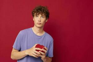 red-haired curly guy a box in the form of a heart in his hands red background photo