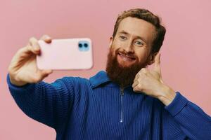 Portrait of a redheaded man with phone in hand taking selfies and photos on his phone with a smile on a pink background, blogger