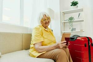 Happy senior woman with passport and travel ticket packed a red suitcase, vacation and health care. Smiling old woman joyfully sitting on the sofa before the trip raised her hands up in joy. photo