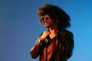Portrait of a stylish man with curly hair with glasses and headphones on a blue background multinational, colored light, black leather jacket trend, modern concept. photo