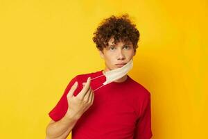 retrato de un joven Rizado hombre vistiendo un rojo camiseta médico máscara en el cara posando amarillo antecedentes inalterado foto