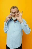 Photo of retired old man in a blue shirt and glasses talking on the phone yellow background