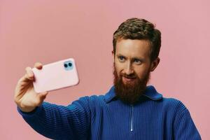 Portrait of a redheaded man with phone in hand taking selfies and photos on his phone with a smile on a pink background, blogger