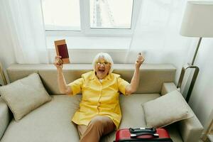 Happy senior woman with passport and travel ticket packed a red suitcase, vacation and health care. Smiling old woman joyfully sitting on the sofa before the trip raised her hands up in joy. photo