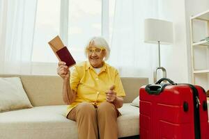 contento mayor mujer con pasaporte y viaje boleto lleno un rojo maleta, vacaciones y salud cuidado. sonriente antiguo mujer alegremente sentado en el sofá antes de el viaje elevado su manos arriba en alegría. foto