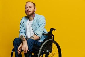 un hombre en un silla de ruedas sonrisa y felicidad, pulgar arriba, con tatuajes en su manos se sienta en un amarillo estudio fondo, el concepto de salud un persona con discapacidades foto