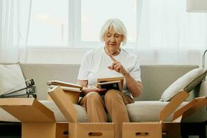 elderly woman sits on a sofa at home with boxes. collecting things with memories albums with photos and photo frames moving to a new place cleaning things and a happy smile. Lifestyle retirement.