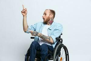 A man in a wheelchair and pointing a finger at, copy space, with tattoos on his hands sits on a gray studio background, health concept a person with disabilities, a real person photo