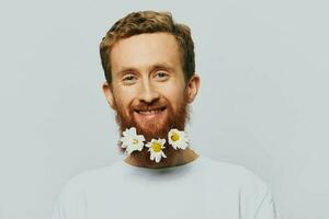 Portrait of a funny man in a white T-shirt with flowers daisies in his beard on a white isolated background, copy place. Holiday concept and congratulations. photo