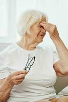 mayor mujer grave ojo dolor sentado en el sofá, salud problemas en antiguo edad, pobre calidad de vida. un abuela con gris pelo sostiene en a su dolorido ojos con lentes en su mano. foto