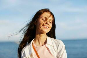 A girl with her eyes closed in the sun against an ocean backdrop smiling with teeth, flying hair, tanned skin, relaxing, traveling to the ocean. photo