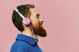 Portrait of a redheaded man wearing headphones smiling and dancing, listening to music on a pink background. A hipster with a beard. photo