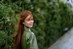 mujer con un hermosa sonrisa posando en contra un verde bambú en primavera, rojo volador pelo en el Ventoso clima, el concepto de estilo y Moda en el ciudad foto