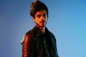 Portrait of a stylish man with curly hair on a blue background multinational, colored light, black leather jacket trend, modern concept. photo